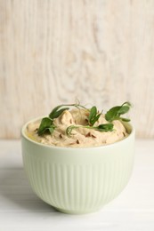 Bowl of tasty hummus with pea leaves on white wooden table, space for text