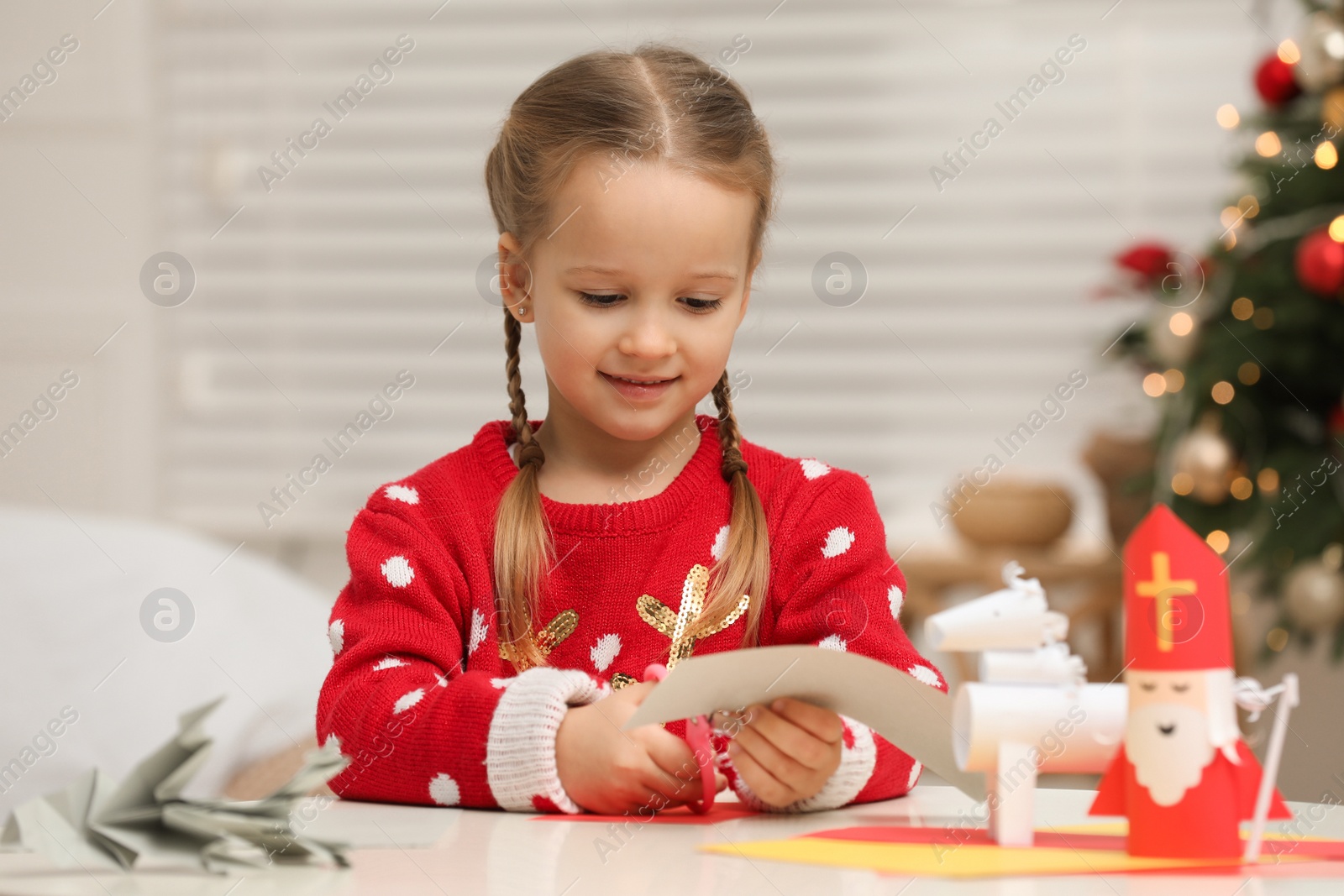 Photo of Cute little girl cutting paper at table with Saint Nicholas toy indoors