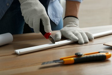 Photo of Worker cutting foam crown molding with utility knife at wooden table, closeup