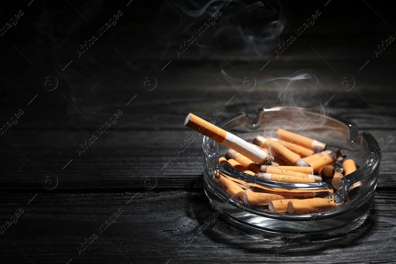 Photo of Smoldering cigarette in glass ashtray on dark wooden table. Space for text