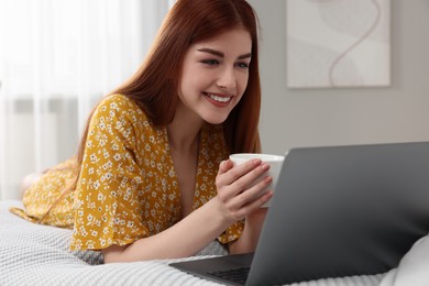 Photo of Happy woman with cup of drink and laptop on bed in bedroom