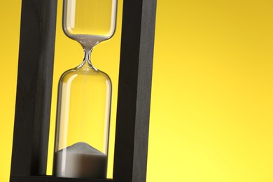 Photo of Hourglass with flowing sand against yellow background, closeup. Space for text