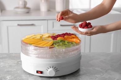 Photo of Woman putting raspberry into fruit dehydrator machine at grey marble table in kitchen, closeup