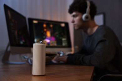 Photo of Young man with energy drink playing video game at wooden desk indoors, focus on can