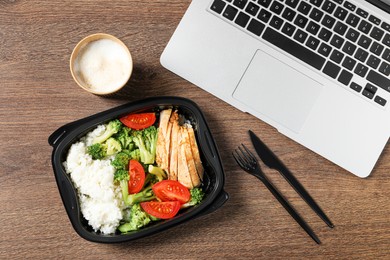 Photo of Container with tasty food, laptop, cutlery and cup of coffee on wooden table, flat lay. Business lunch
