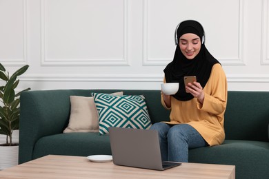 Muslim woman in hijab with cup of coffee and smartphone using laptop on sofa indoors. Space for text