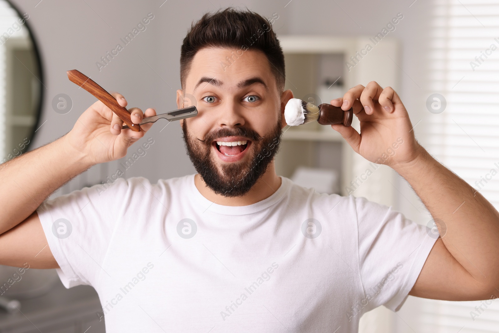 Photo of Handsome young man holding blade and shaving brush with foam in bathroom