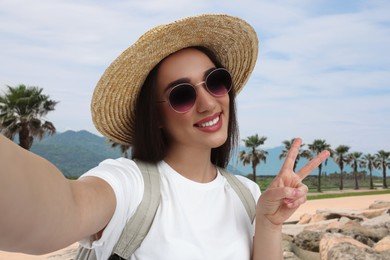 Image of Smiling young woman in sunglasses and straw hat taking selfie and showing peace sign outdoors