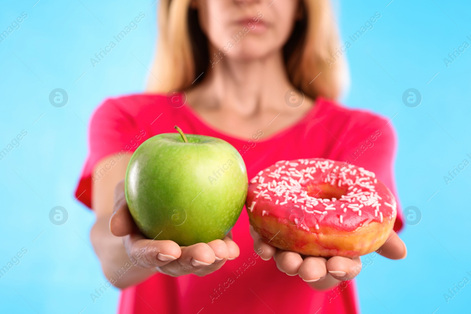 Photo of Woman choosing between doughnut and fresh apple on light blue background, closeup