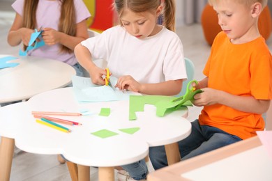 Photo of Cute little children cutting color paper with scissors at desks in kindergarten. Playtime activities