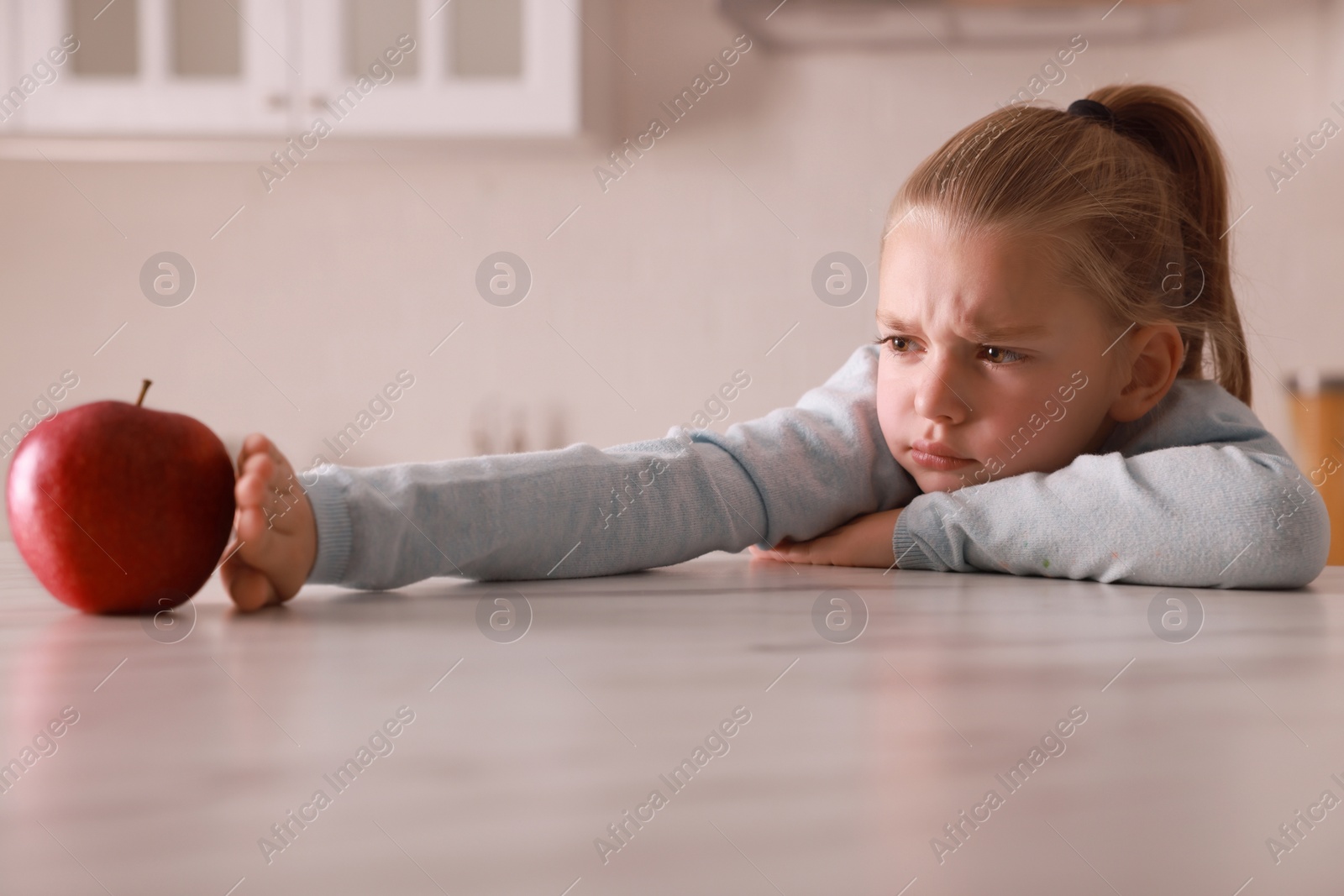 Photo of Cute little girl refusing to eat apple in kitchen