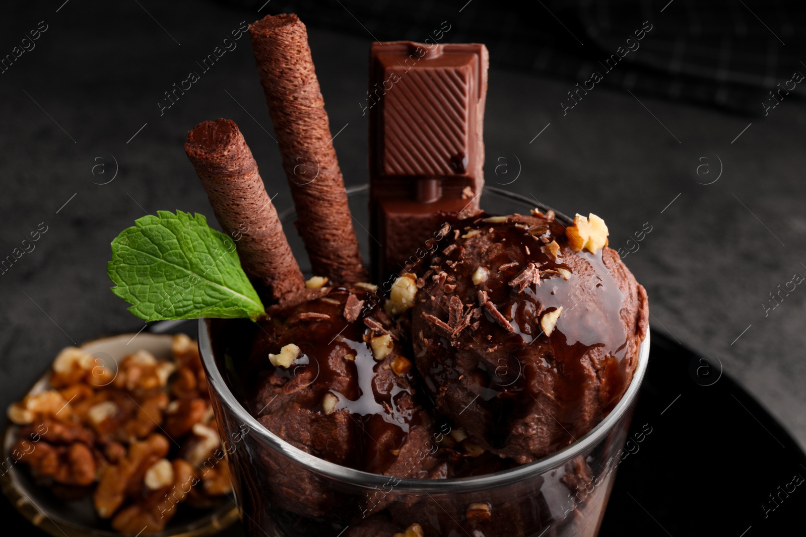 Photo of Tasty chocolate ice cream with sauce, nuts and wafer rolls in glass dessert bowl on table, closeup