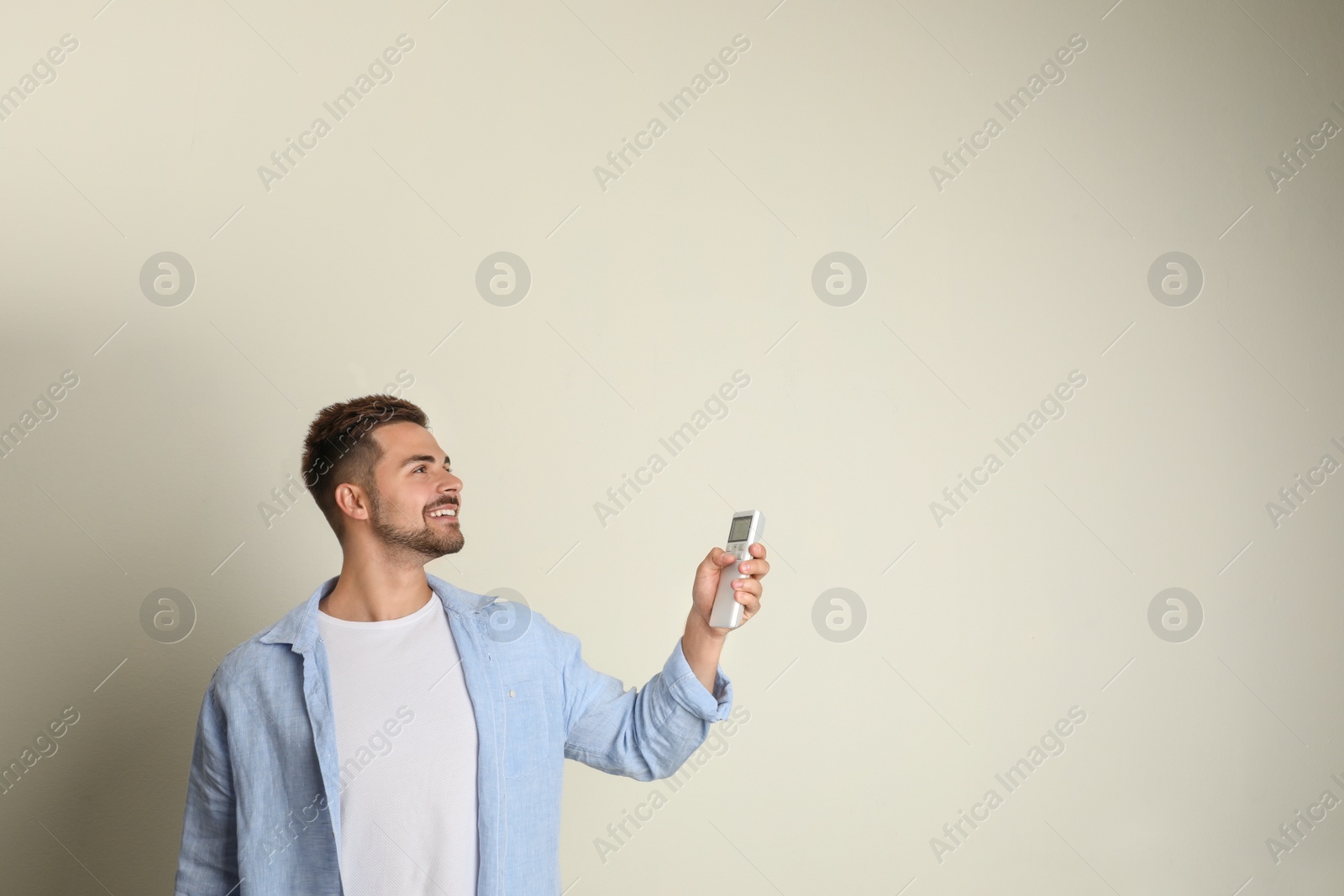 Photo of Happy young man operating air conditioner with remote control on beige background. Space for text