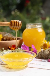 Photo of Delicious honey flowing down from dipper into bowl on white wooden table in garden