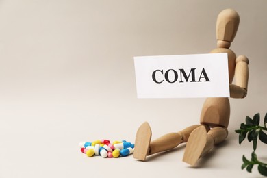 Human mannequin holding card with word Coma and pills on beige background. space for text