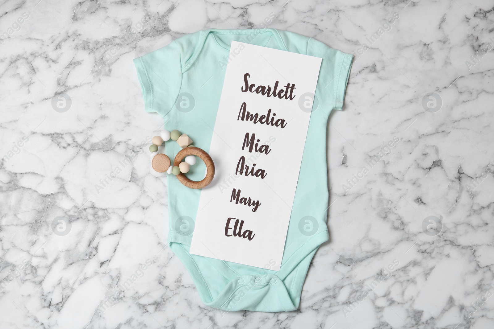 Photo of Bodysuit with list of baby names and toy on white marble background, top view