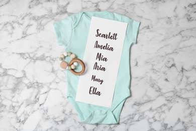 Bodysuit with list of baby names and toy on white marble background, top view