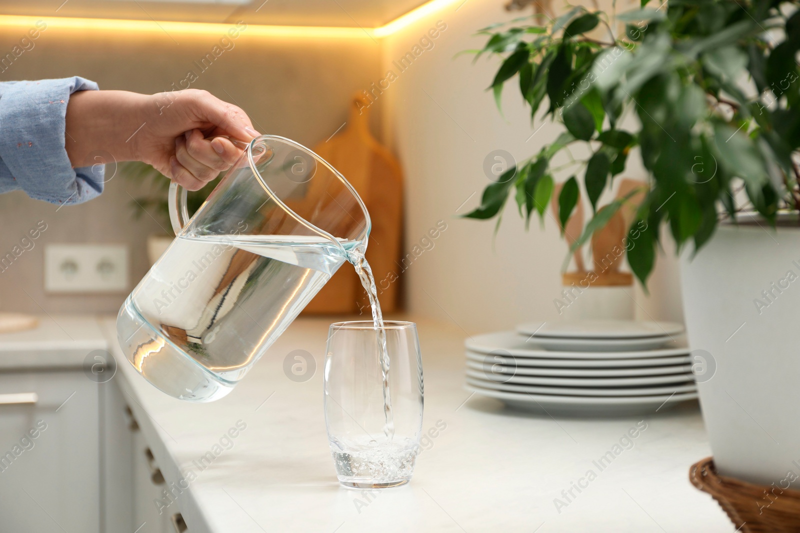 Photo of Woman pouring water from jug into glass at white table in kitchen, closeup