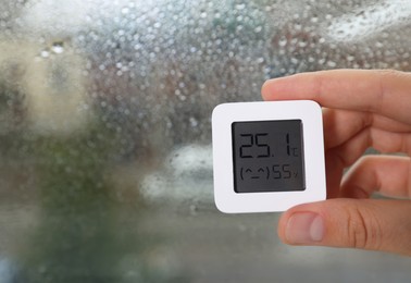 Photo of Woman holding digital hygrometer with thermometer near window on rainy day, closeup. Space for text