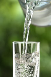 Photo of Pouring water from jug into glass on blurred green background, closeup