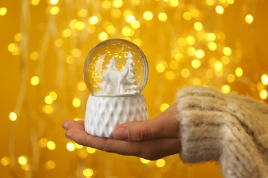 Woman holding snow globe with fir trees and deer against festive lights, closeup