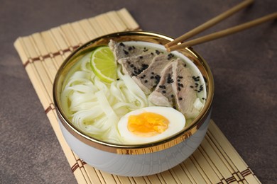 Photo of Bowl of delicious rice noodle soup with meat and egg on brown table, closeup