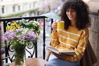 Young woman with cup of drink reading book near beautiful flowers on table at balcony