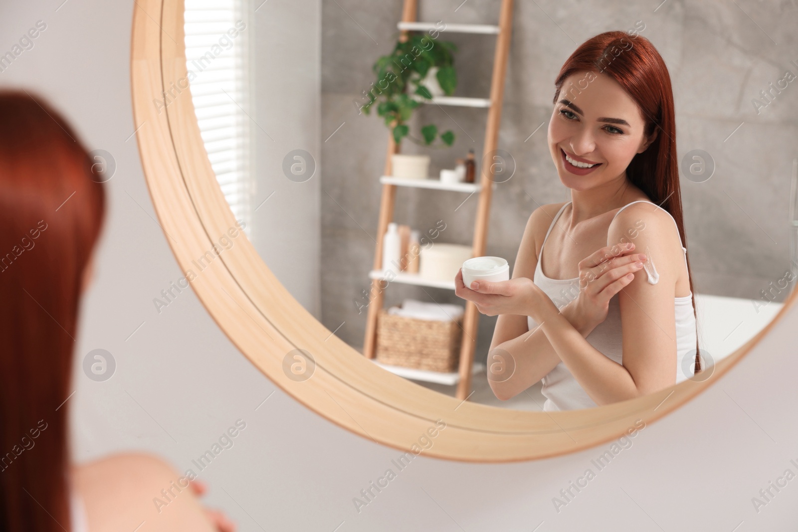 Photo of Beautiful young woman applying body cream onto shoulder near mirror in bathroom