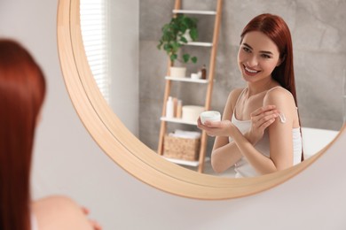 Beautiful young woman applying body cream onto shoulder near mirror in bathroom