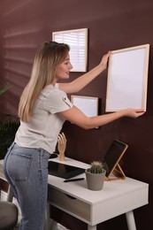 Young woman hanging empty frame on brown wall indoors. Mockup for design