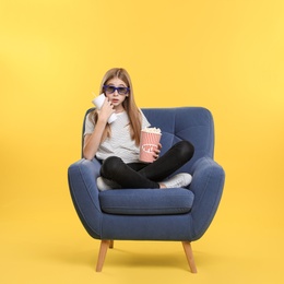 Emotional teenage girl with 3D glasses, popcorn and beverage sitting in armchair during cinema show on color background