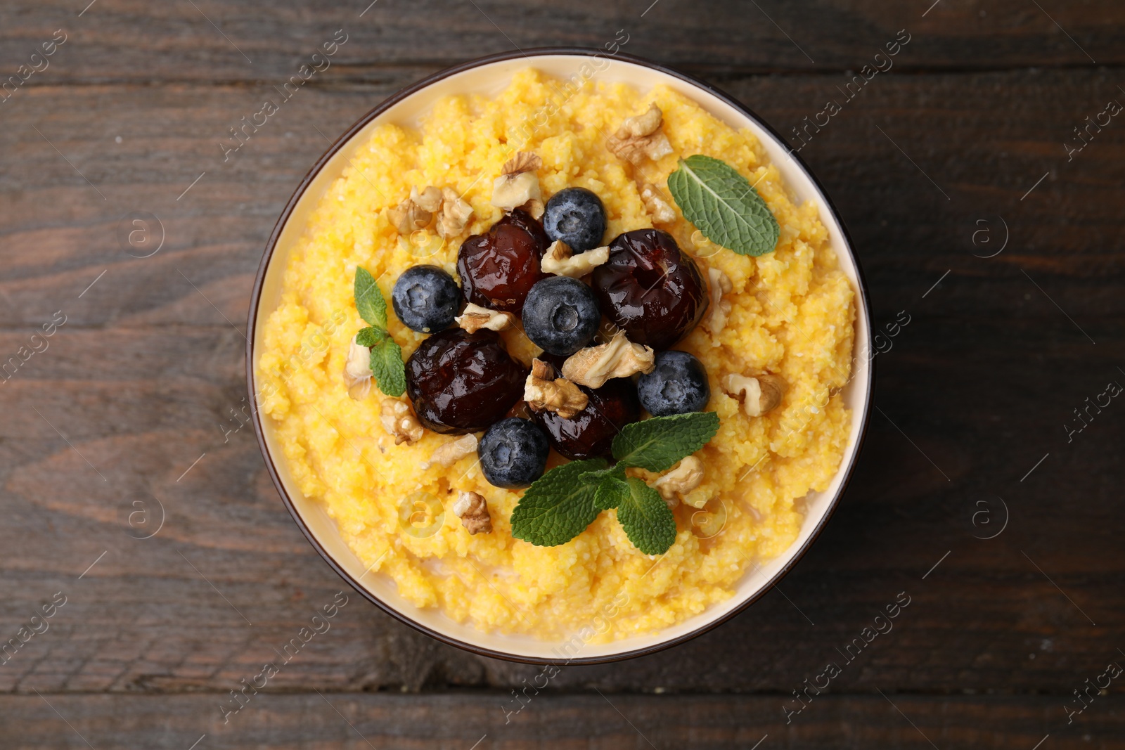 Photo of Tasty cornmeal with blueberries, dates, walnuts and mint in bowl on wooden table, top view