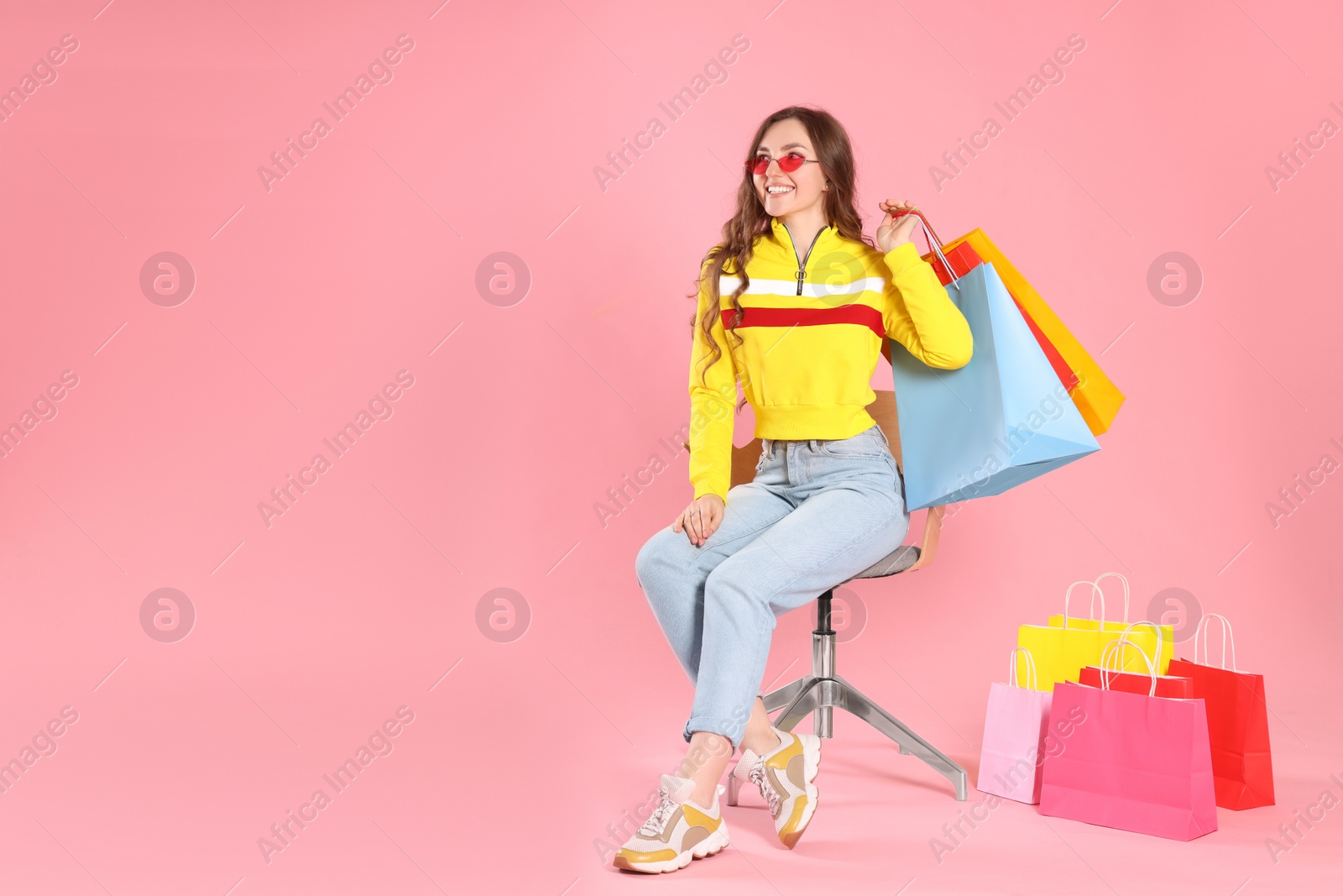 Photo of Happy woman in stylish sunglasses holding many colorful shopping bags on armchair against pink background