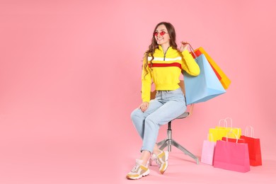 Happy woman in stylish sunglasses holding many colorful shopping bags on armchair against pink background