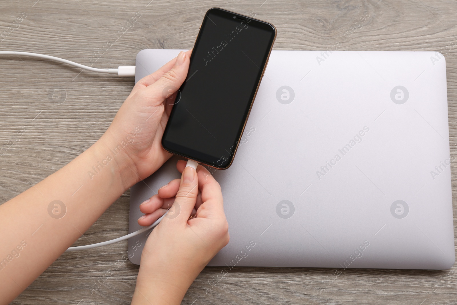 Photo of Woman connecting smartphone with charge cable to laptop on wooden table, top view. Space for text