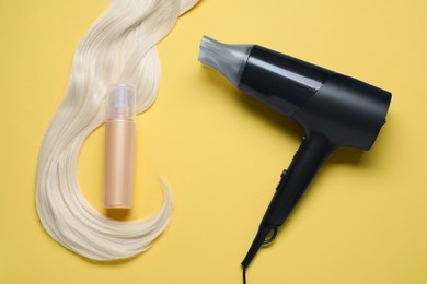 Spray bottle with thermal protection, hairdryer and lock of blonde hair on yellow background, flat lay