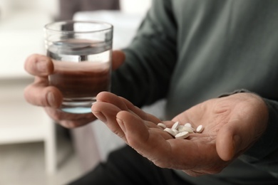 Senior man holding pills and glass of water indoors, closeup