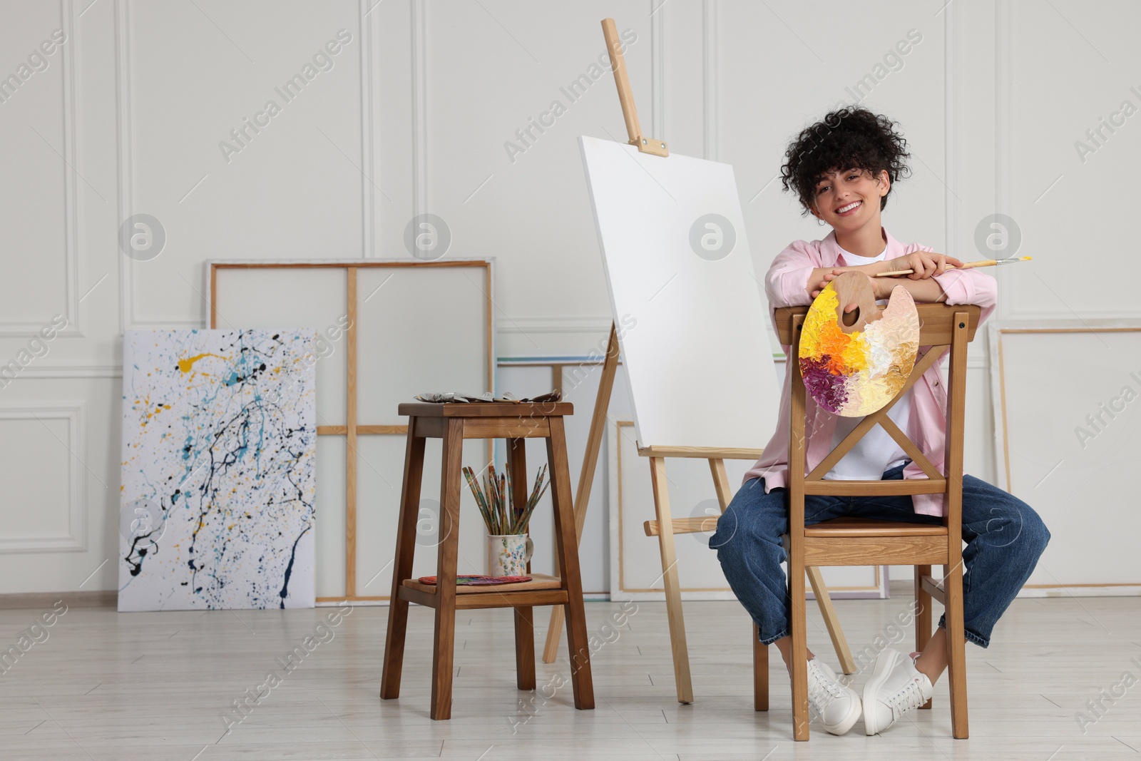 Photo of Young woman holding brush and artist`s palette near easel with canvas