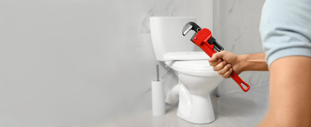 Image of Closeup view of professional plumber holding pipe wrench near toilet bowl in bathroom, space for text. Banner design