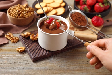 Photo of Woman dipping strawberry into fondue pot with milk chocolate at wooden table