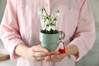 Photo of Woman holding pot with beautiful snowdrops and traditional martisor indoors, closeup. Symbol of first spring day