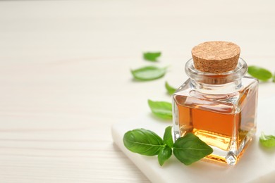 Photo of Bottle of essential basil oil and fresh leaves on white wooden table, space for text