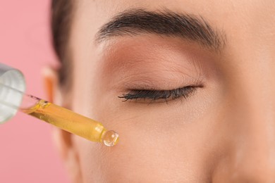 Young woman applying serum onto her face on pink background, closeup