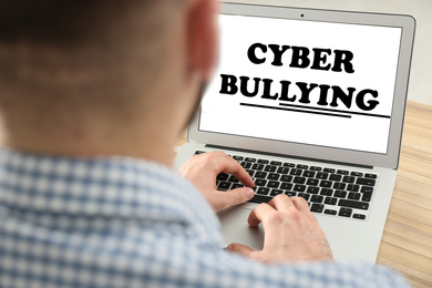 Young man using laptop at wooden table, closeup. Cyber bullying