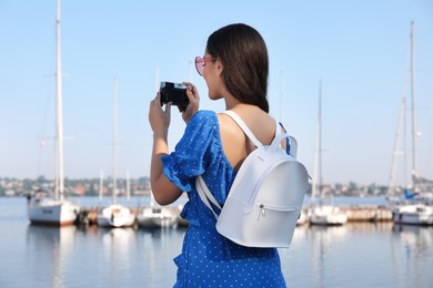 Young woman with stylish backpack and camera near river on sunny day, back view