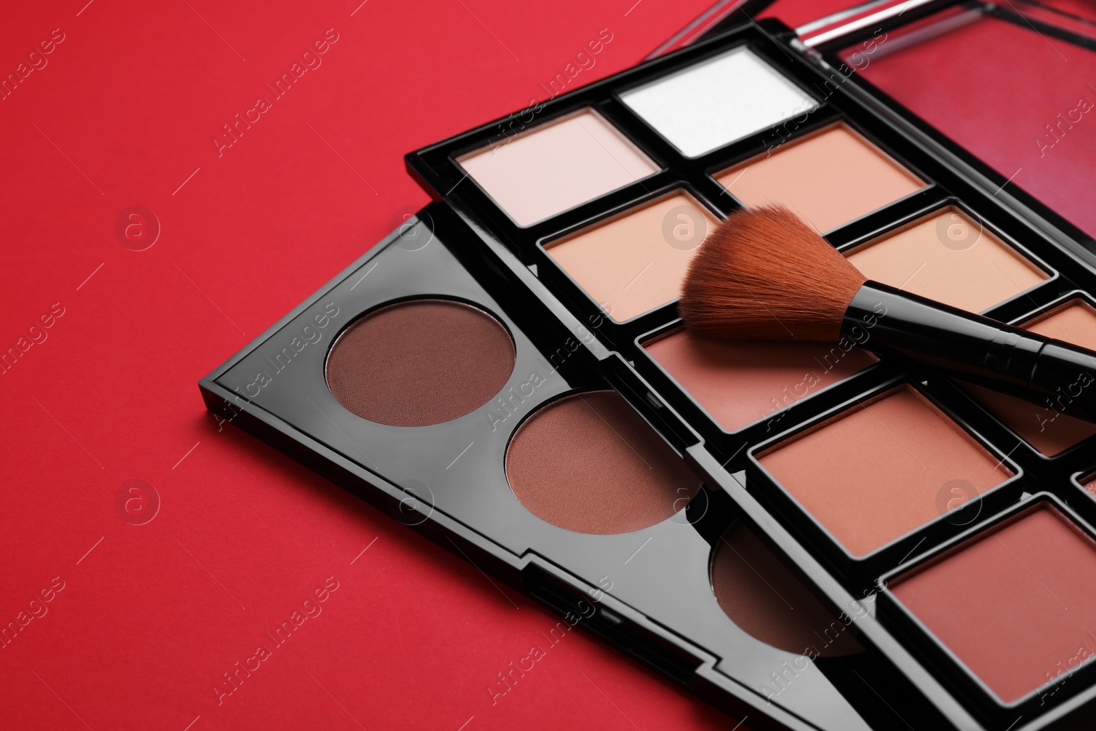 Photo of Colorful contouring palettes with brush on red background, closeup. Professional cosmetic product