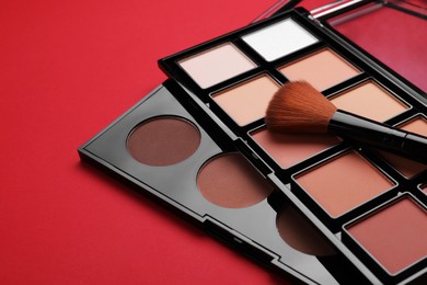 Photo of Colorful contouring palettes with brush on red background, closeup. Professional cosmetic product