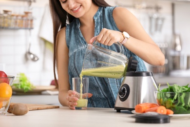 Photo of Young woman pouring tasty healthy smoothie into glass at table in kitchen