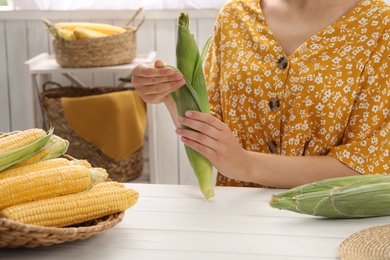 Photo of Woman husking corn cob at white wooden table, closeup