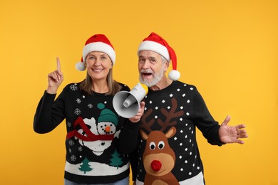 Photo of Senior couple in Christmas sweaters and Santa hats shouting in megaphone on orange background